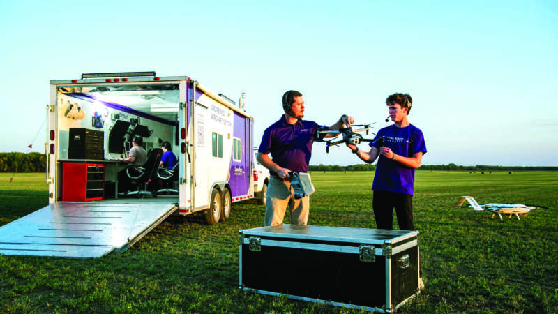 two students holding an unmanned aircraft system in a field near a trailor