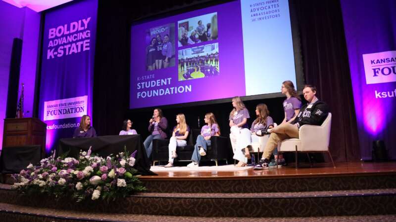 students sitting on chairs on a stage talking with a presentation in the background