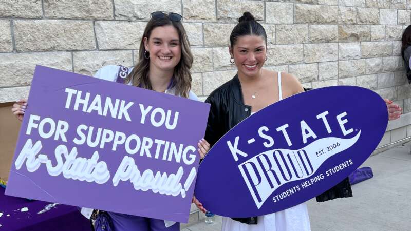 two female students holding signs reading "Thank you for supporting K-State Proud" and "K-State Proud"